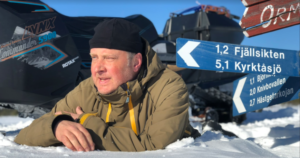En bild på Anders som ligger på magen i en snödriva och kisar mot solen. I bakgrunden syns olika skyltar med distanser till olika platser och en snöscooter.